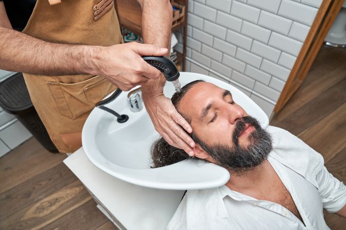 Crop barber washing hair of bearded man