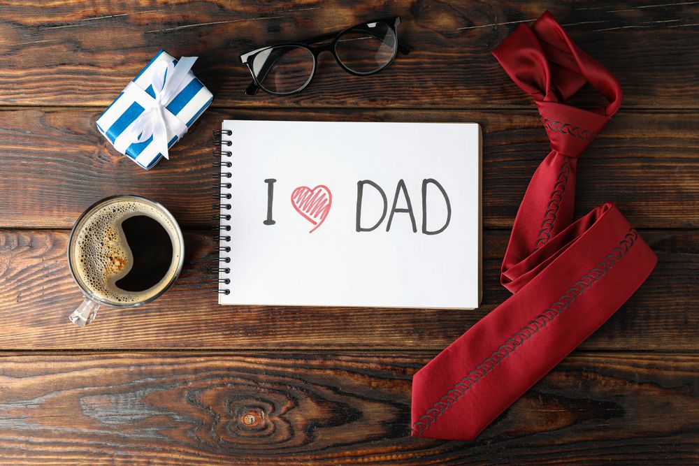Notebook with inscription I love DAD, cup of coffee, glasses, gift box and tie on wooden background, space for text and top view