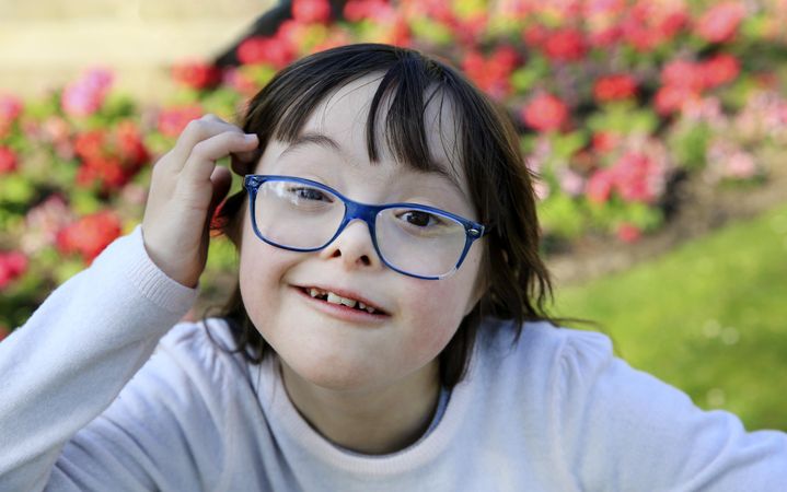 Adorable little girl with blue eyeglasses smiling and looking at camera with flowers behind