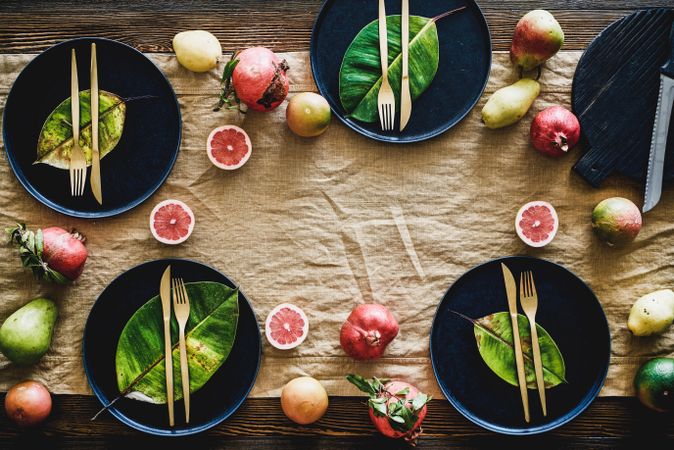Fresh table setting with leaves on dark plates, scattered fruit on brown table cloth, copy space