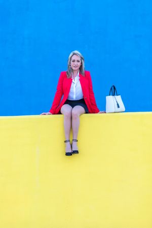 Woman in red coat sitting on bright yellow wall with blue in background