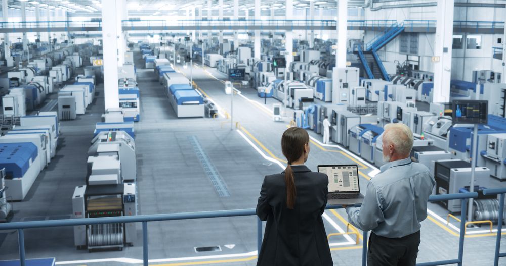 Male Engineer and Female Project Manager Standing on a Platform, Talking, Using Laptop Computer and Monitoring Production at a Modern Automated Electronics Manufacture with AI Robotic Arms