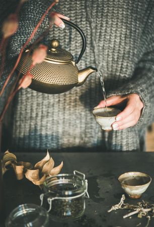 Woman in cozy sweater pouring from traditional Japanese tea set, with dried poppies