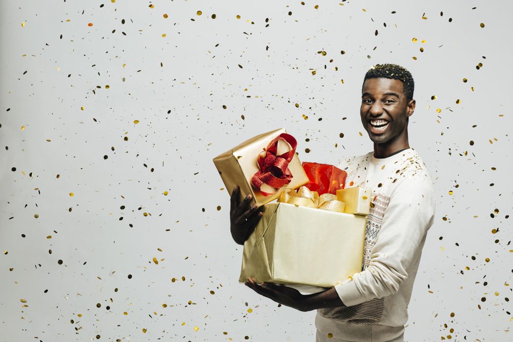 Smiling Black man holding pile of wrapped presents in both arms as glitter falls