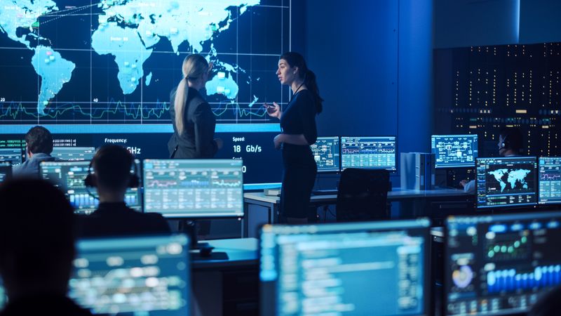 In a Cutting-edge Control Room, Diverse Professionals Engage in Dynamic Discussions, Analyzing Global Data on Multiple Screens to Enhance Cybersecurity Measures and Strategic Operations.