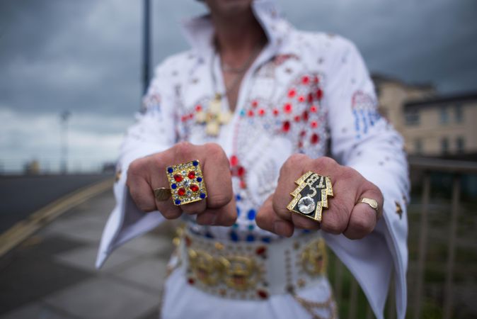 Elvis impersonator showing off flashy rings on sidewalk with blurry, cloudy background