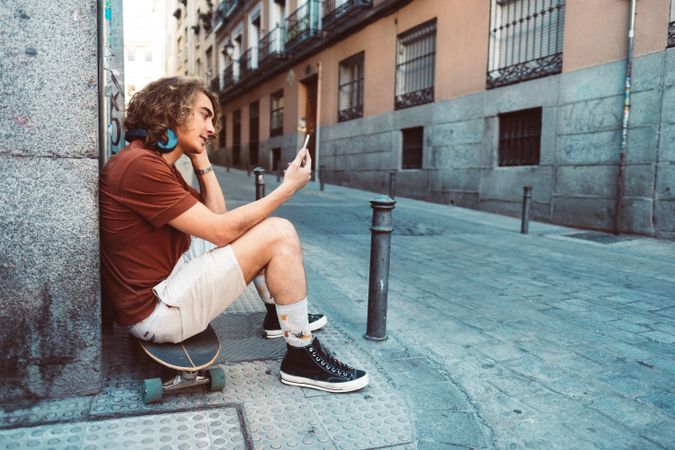 Relaxed man in headphones using smartphone while resting on longboard on street