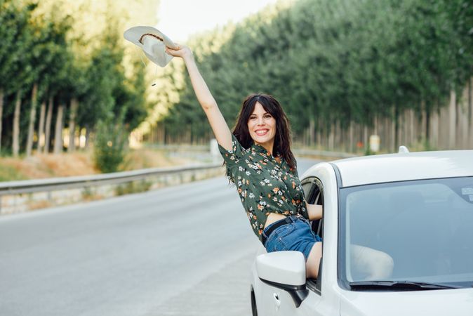 Smiling brunette female waving straw hat outside passenger window