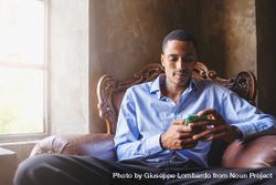 A Black man in a shirt types on his smartphone sitting in a comfortable armchair bE9pW7