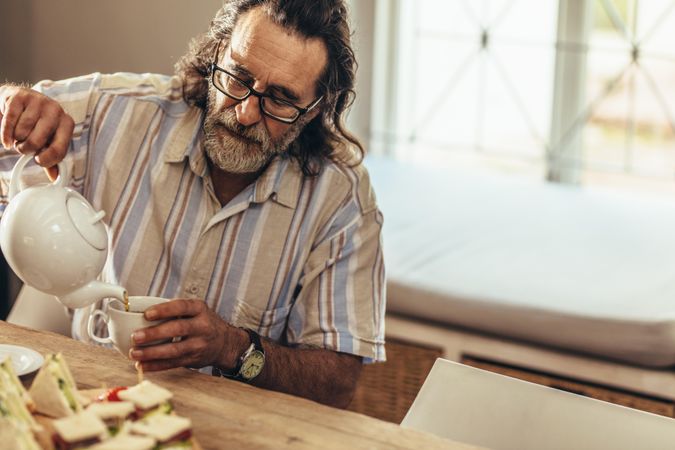 Man with beard pouring tea in from teapot to cup while sitting at table indoors
