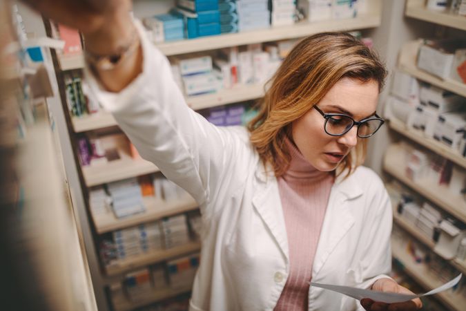 Chemist looking at prescription while searching prescribed drug on shelves in pharmacy