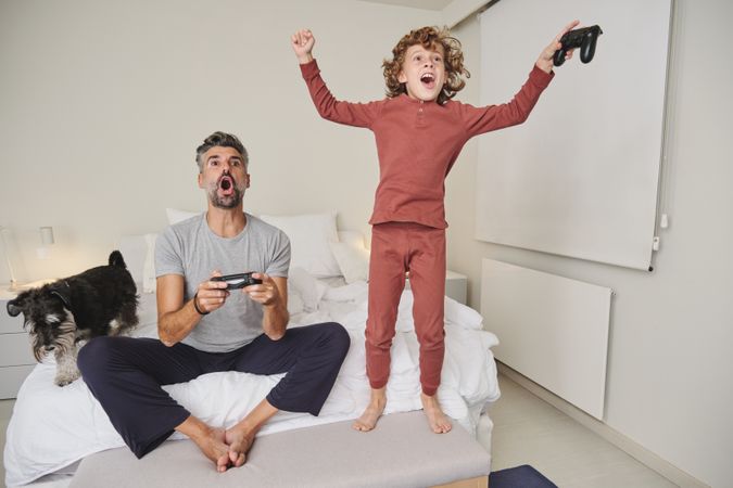 Excited father and son playing videogame in bedroom with dog