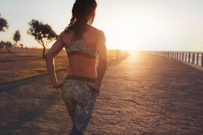 Rear view image of fitness woman in sportswear walking on a seaside promenade at sunset