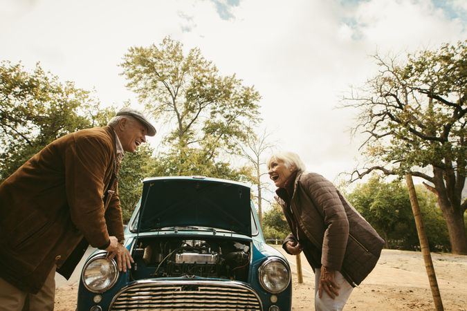 Mature couple laughing in front of their broken down car with the hood open