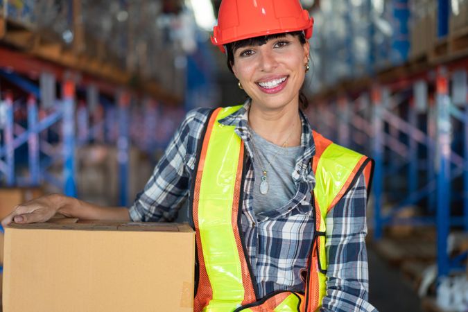 Woman in hard hat smiling and carrying in box in shipping center