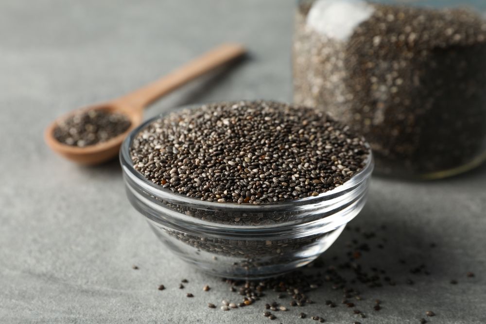 Glass bowl, jar and wood spoon with chia on grey background, closeup