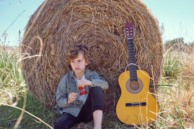 Upset kid sitting near guitar and leaning on hay bale