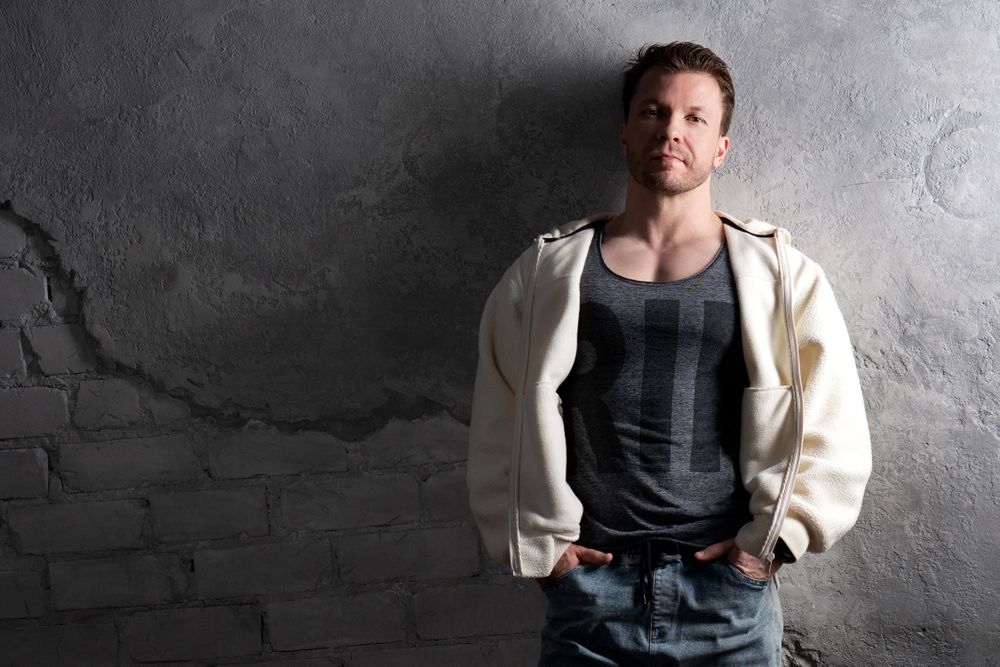 Man in fleece hoodie and jeans leaning against wall.