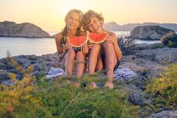 Cheerful kids sitting on rocky seashore with watermelon