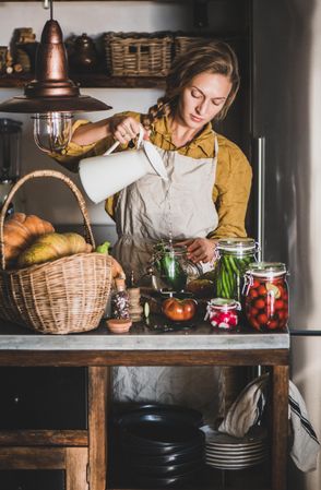 Young woman in rustic kitchen pouring vinegar into jar of cucumbers for pickling
