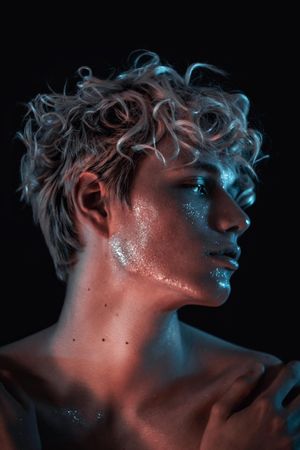 Side view portrait of topless blonde man with UV paint on his face against dark background