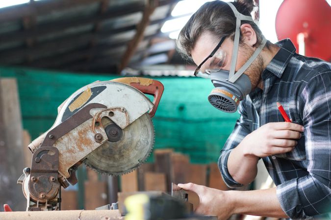 Male carpenter wearing protective mask using electric circular saw cutting wood board at workshop