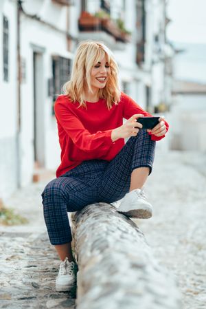 Happy blonde female with red shirt texting while sitting outdoors, vertical