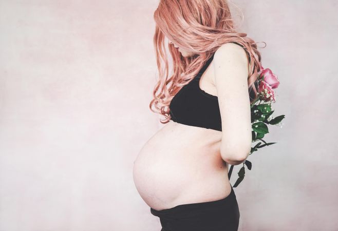 Side view of pregnant woman with pink hair wearing crop top holding pink rose bouquet