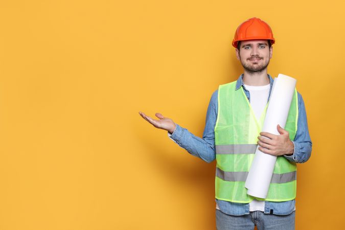 Engineer in helmet with drawing paper in hand on yellow background, space for text