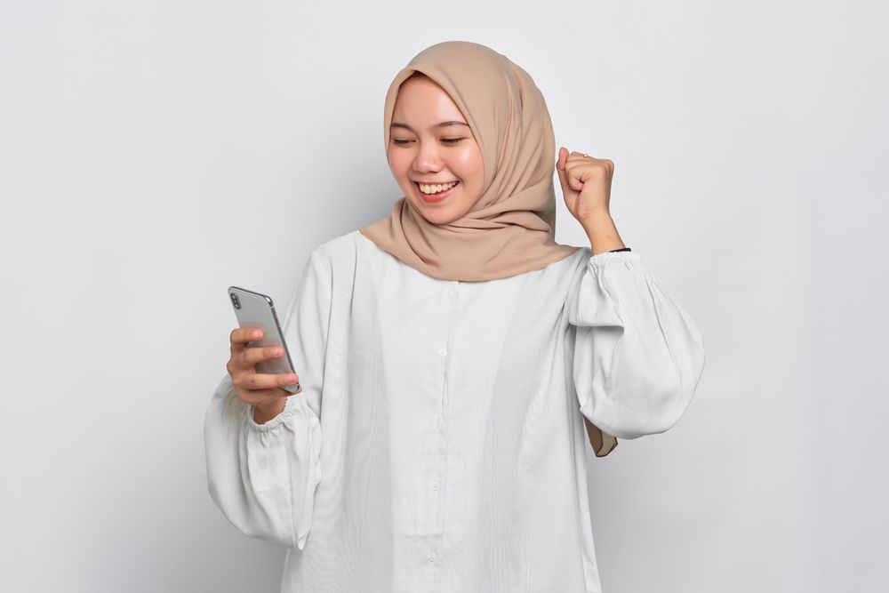Asian female in headscarf receiving happy news on her cell phone