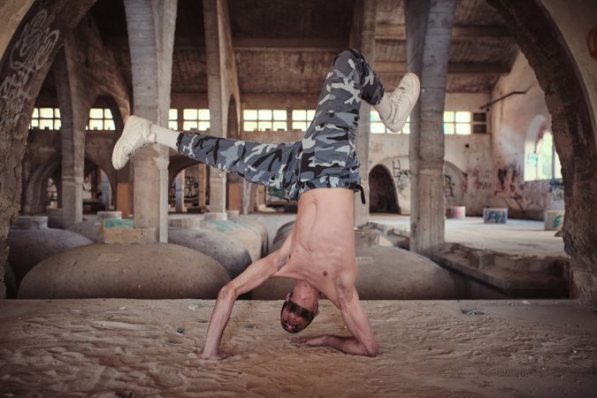 Muscular man performing cricket movement of breakdance in abandoned building