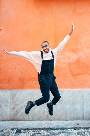 Male in overalls and hoodie smiling and jumping down in front of wall