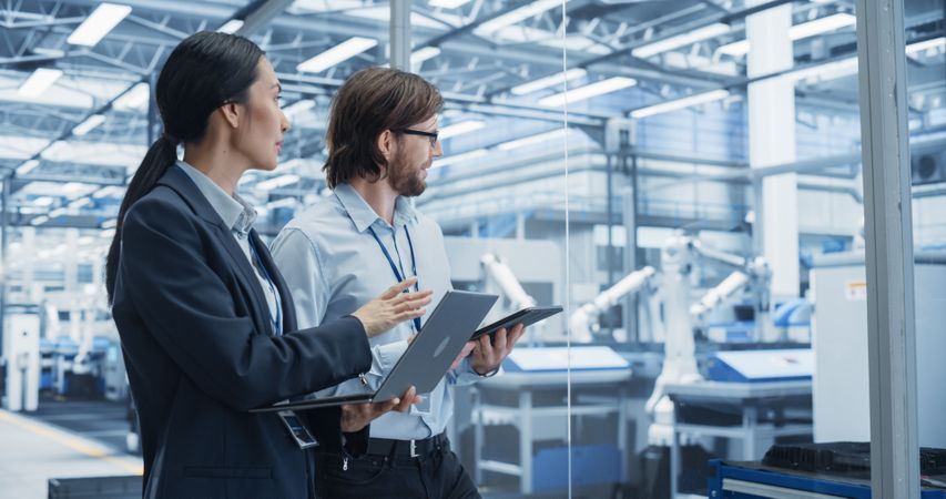 Portrait of Cheerful Engineer and Happy Asian Scientist Using Laptop and Tablet Computers, Talking in a Factory Facility with Robots Producing Modern Electronic Components for Different Industries