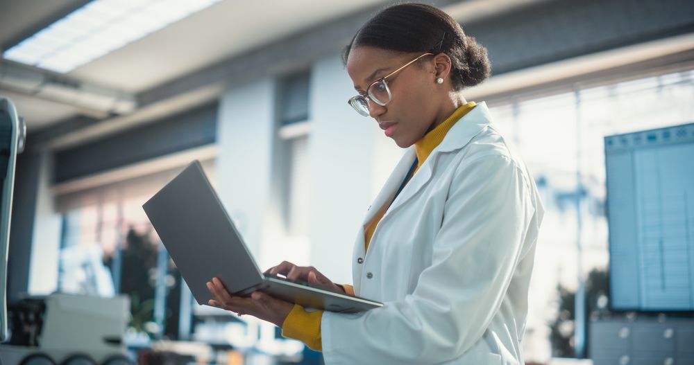 Portrait of Young Black Female Specialist in Lab Coat Using Laptop Computer to Test an AI Robotic Prototype. Professional,
