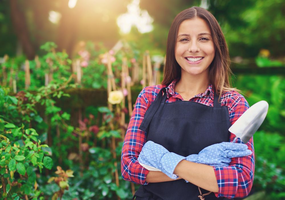 Confident woman pictured in a greenhouse with her arms crossed holding a a trowel
