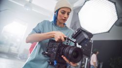 Focused Young Female Cinematographer Adjusts Professional Camera During Shoot, With Lighting Equipment And Colleague In Background, Highlighting Creativity In Film Production And Teamwork.