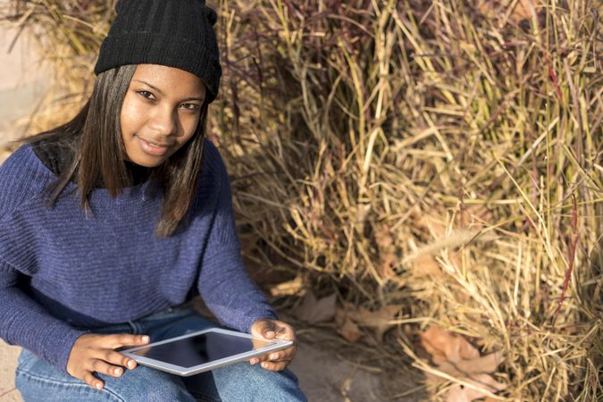 Confident female in hat and sweater sitting in grass with tablet