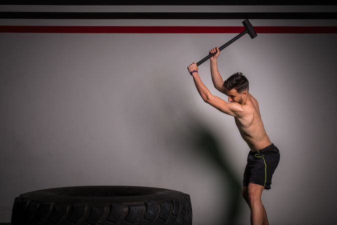Shirtless guy concentrated in sportswear with hammer hitting on big tire in gym