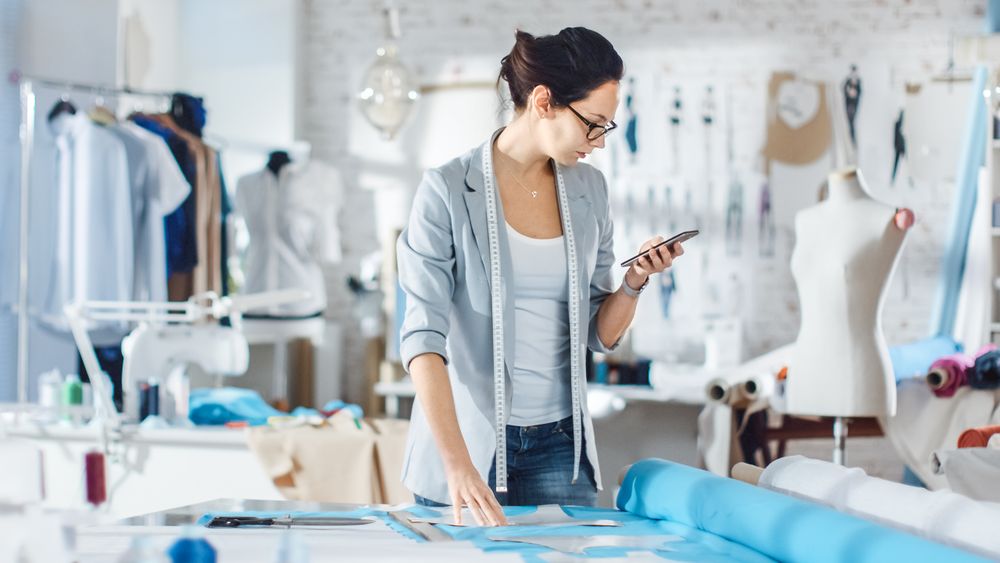 Brunette fashion, designer,  Lays Out Templates on the fabrics and Consults Her Smartphone.
