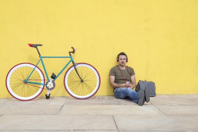 Male sitting in front of yellow wall next to bike, and bag listening to music on smartphone