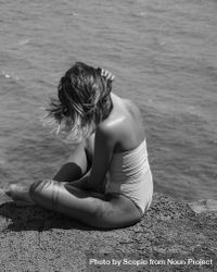 Back view of blonde woman in one piece swimsuit sitting by shoreline in grayscale 5oOwm5