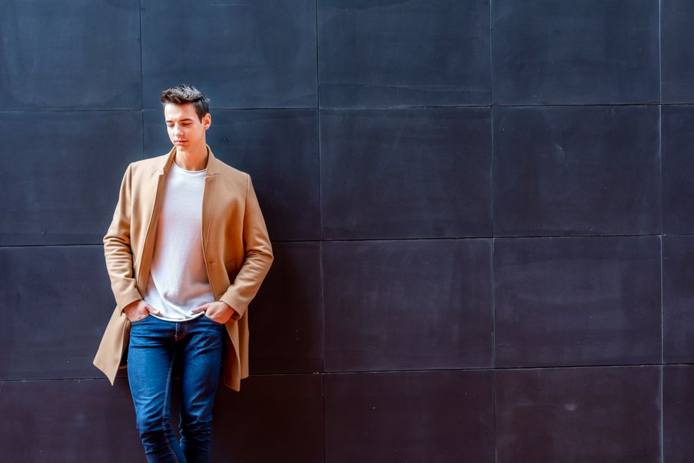 Male in camel coat looking down leaning against dark wall