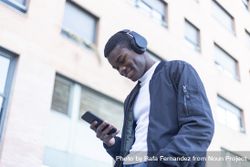Smiling Black man leaning outside, wearing headphones and looking down at his phone 5zr7mX