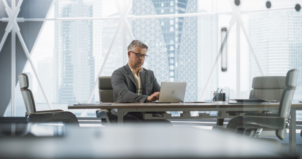 Portrait of Successful Businessman Working on Laptop Computer in His Big City Corner Office.
