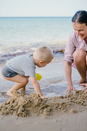 Toddler and mother playing with sand on beach against blurred seascape in sunny day