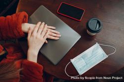 Top view of woman in red jacket sitting at a table with laptop and phone and cup of coffee along with facemask 5rAY30