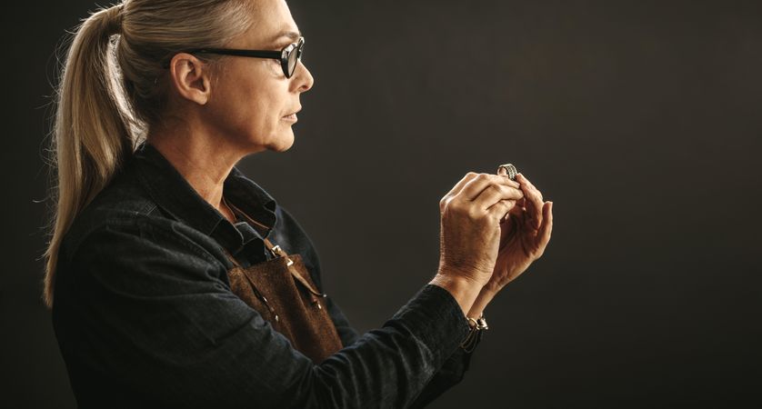 Woman jeweler wearing eyeglasses and apron looking at  ring against gray background