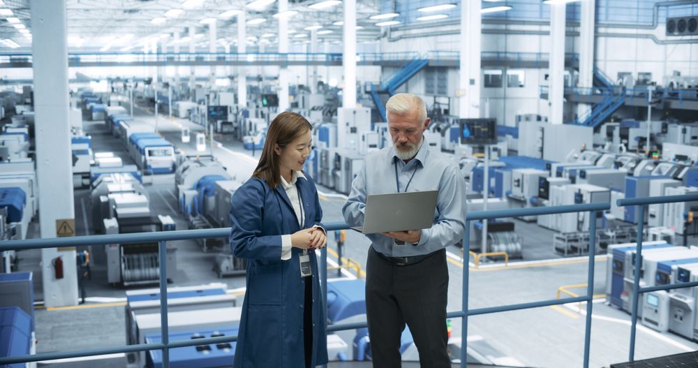 Factory Managers Working on Laptop at a Modern Manufacture with Autonomous Robot Hands. Female and Male Specialists Monitoring Technical Data on a Computer, Analyzing Reports and Planning Updates