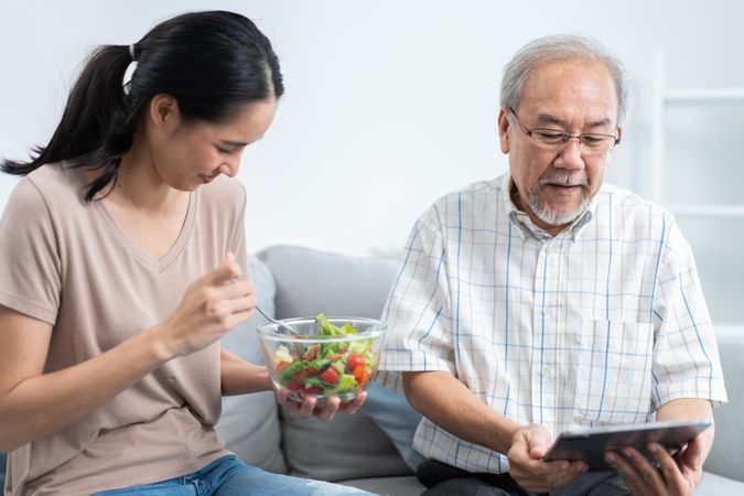 Older Asian male using digital as his daughter serves salad