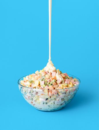 Pouring mayo over oliveir salad, minimalist on a blue background